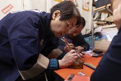 A woman wearing a dark blue jacket and glasses uses a small wooden tool to work on a hand-carved seal while another person looks at a smartphone.