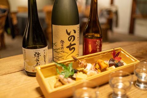 Kitakata Day Tour: Sake Tasting & Food Pairing Experience Three bottles of sake and a bento box with assorted food items and two small sake glasses are arranged on a wooden table.