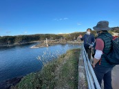 Two people stand on a walkway overlooking a body of water with a rocky coastline and some distant structures.