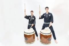 Two performers in traditional Japanese attire play large drums with wooden sticks against a white backdrop.