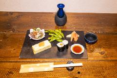 A Japanese appetizer platter sits on a wooden table, featuring edamame, sushi rolls, rolled omelet, and a sake carafe and cup.