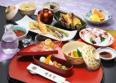 An elegant Japanese meal arrangement, featuring a variety of dishes like tempura, sashimi, fruits, and sake, beautifully presented on a red lacquered tray against a backdrop of textured fabrics.