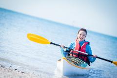 A smiling woman in a kayak holds a paddle, wearing a life vest, enjoying a sunny day on the water.