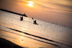 Two kayakers paddle on water at sunset, with sailboats and a breakwater with a building visible in the distance.