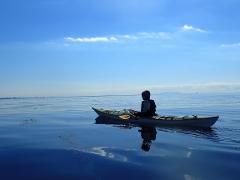 A serene image of a kayaker peacefully gliding across calm, reflective water under a clear blue sky with scattered clouds.