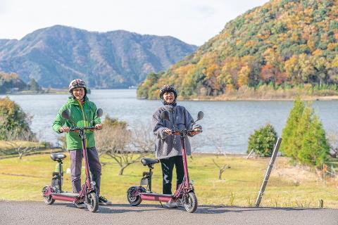 Mikata Lakes Fukui Tour: Kickboard Adventure & Varve Discovery Two people in helmets pose with their electric scooters in front of a scenic lake and mountains, with autumn foliage in the background.