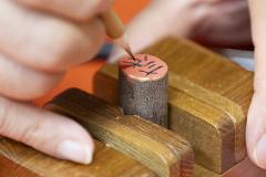 A close-up shot shows hands using a brush to paint characters onto a wooden cylindrical seal being held in a carving vise.