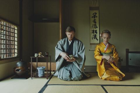 A man and a woman in traditional Japanese clothing kneel on tatami mats, holding tea bowls in a serene, ritualistic setting.