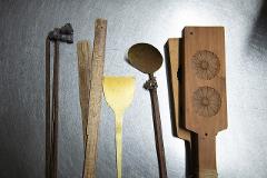 A flat lay of traditional Japanese sweets making tools, including wooden molds with flower imprints, metal spatulas, and metal rods with attachments, rests on a brushed metal surface.