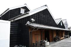 A traditional Japanese building with dark wooden walls and white-tiled roofs stands under a bright sky, with vintage machinery and a wooden bench outside.