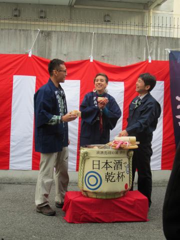 Three people stand around a sake barrel, holding small wooden cups in front of a red and white striped backdrop, participating in a Japanese ceremony.