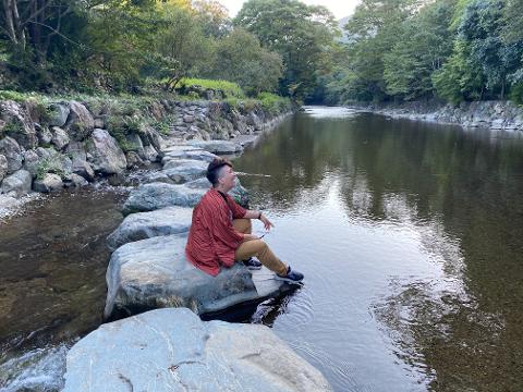 Ise Sacred River EV Ride – 90 Min Scenic Tour from Senokuniya A person sits on stepping stones in a clear river, surrounded by trees and rocks.