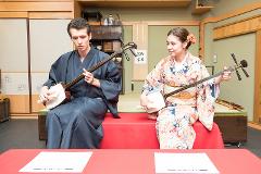 Play a Traditional Japanese String Instrument -- "Shamisen" A man and a woman in traditional Japanese attire play the shamisen, a three-stringed instrument, while sitting on a red cushion.