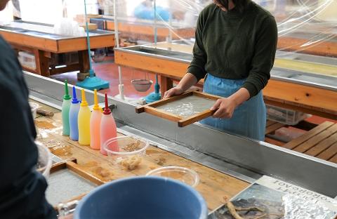 A person in a green sweater and blue apron uses a wooden frame to make Japanese Washi paper, with colorful squeeze bottles and craft materials nearby.