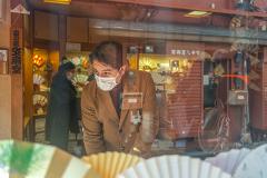 A man wearing a mask and a brown coat looks at a display of fans in a shop window.
