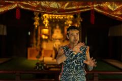 A man in traditional Japanese attire performs a dance with expressive hand gestures in a dimly lit temple adorned with red and gold decorations.