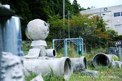 A concrete sculpture resembling a robot is placed in a grassy outdoor area with a school building in the background.