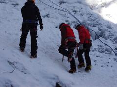 People in ice climbing gear ascend a snow-covered slope with ice formations visible in the background.