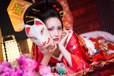 Eine elegante Asiatin in einem roten Kimono und mit Geisha-Frisur hält eine Fuchsmaske vor ihr Gesicht, während sie vor einem gemusterten Hintergrund liegt.