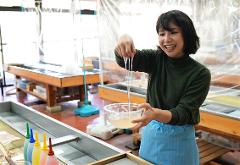 A smiling woman in a green sweater and blue apron is holding a bowl of translucent liquid with three long, clear rods suspended in it.