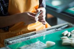 A person wearing an orange apron delicately works with small, white objects, possibly feathers or fibers, on a green and silver work surface.