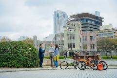 Three people stand talking next to a bush with three bikes parked nearby in front of the Hiroshima Peace Memorial, under a cloudy sky.