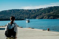 A woman with a backpack stands on a concrete pier, looking out at a boat in the blue sea with a forested island in the background.