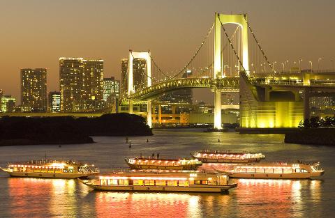This image depicts Tokyo's Rainbow Bridge illuminated at night, with several illuminated boats on the water in the foreground and buildings in the background.