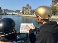 Two people wearing helmets are looking at a photo of a building while standing near a river with the Atomic Bomb Dome in the background.