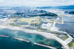 An aerial view captures a coastal area featuring a modern park with water features, a marina, a city in the background, and a bridge spanning a river.