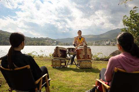 Sacred Drumming Day Tour at Lake Shirakaba, Nagano – Japan 2025 A man plays traditional Japanese drums by a lake while two people watch from chairs in the foreground.