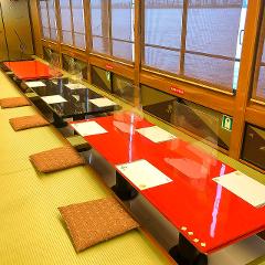 A high-angle shot showcases two long tables, one red and one black, each divided by clear plastic barriers. Floor cushions are placed for seating, and the scene is set against a backdrop of large windows with drawn shades.