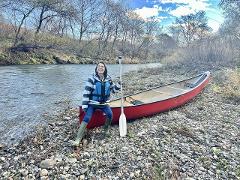 A woman stands proudly by a red canoe on a rocky riverbank, holding a paddle and smiling, ready for a half-day tour on the Shiodomari River.