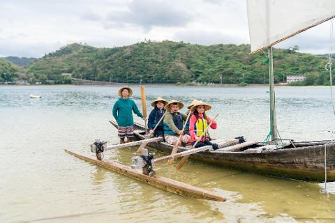 Four people wearing conical hats are on a traditional wooden boat with a sail in a bay.