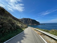 Ishikawa Satoyama Cycling – Wajima Half-Day Nature Experience A paved road curves along a coastline with a steep, green hillside on the left and the ocean on the right, under a bright blue sky.