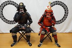 Two individuals in traditional Japanese samurai armor sit on chairs in front of a white backdrop with black decorative circles.