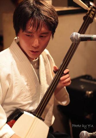 En musiker i en hvid kåbe spiller intenst en traditionel japansk shamisen, placeret foran en mikrofon.