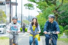 Three people wearing helmets are riding bicycles on a street with trees and buildings in the background.