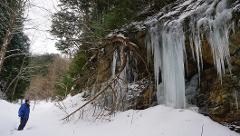 A person stands in a snowy landscape admiring a rock face adorned with icicles in a winter scene.