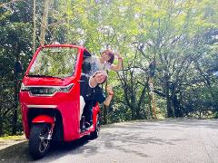 Two smiling people give a thumbs-up while riding in a red electric vehicle through a tree-lined road.