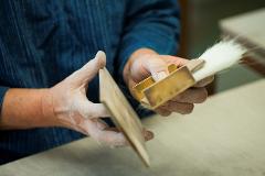 A person in a blue shirt holds a thin wooden object and a brush with a gold handle, suggesting woodworking or cleaning.