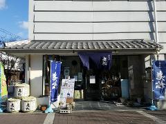 A Japanese sake shop features a display of sake barrels, blue flags with white kanji, and a dark blue banner above the entrance, all advertising the "Oimatsu" (Old Pine) brand.