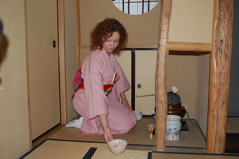A woman in a pink kimono kneels in a traditional Japanese room, preparing tea with a bowl in her hand.