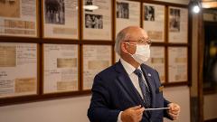 Visit Nikko's Craftsmanship Workshop to Experience Historic Techniques An elderly man wearing a suit, mask, and glasses holds a pointer while standing in front of framed historical documents.