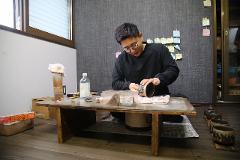 A man in black clothing and glasses is carefully crafting a lacquerware cup on a low table in a room.