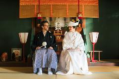 A smiling couple in traditional Japanese wedding attire sits facing each other in a room adorned with red and gold drapery.