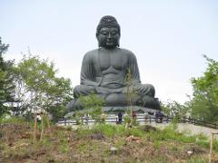 A giant statue of Buddha sits serenely against a pale sky, with lush trees and a few onlookers in the foreground.