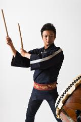 A man in traditional Japanese attire holds drumsticks in front of a taiko drum.