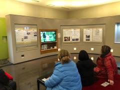 Three people are seated in an exhibition space, watching a screen that displays an aerial view of colorful fish ponds.