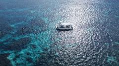 An aerial shot shows a white boat anchored in turquoise waters, surrounded by people snorkeling near a vibrant coral reef.